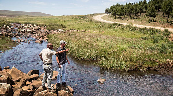 two men in neutral clothing on the edge of a river with a forest in the background. one of the men is pointing towards the forest